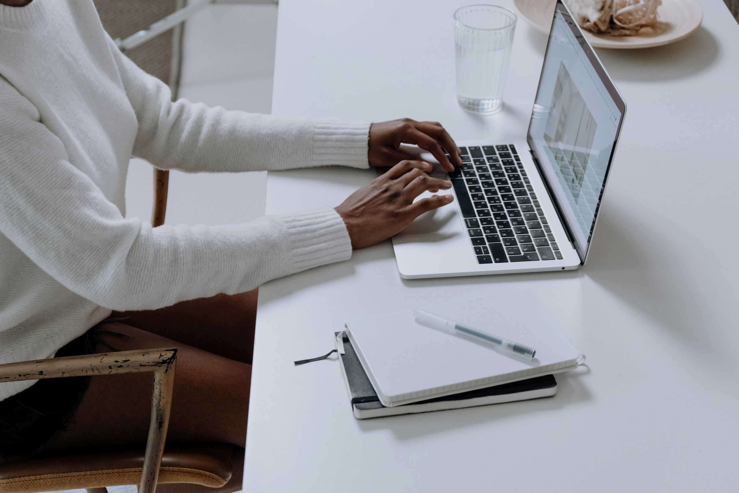 close up of hands typing on a laptop on a white desk next to a note book